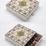 Intricately designed white gift box with layered gold and brown Islamic patterns and a central medallion, opened to reveal assorted premium dates