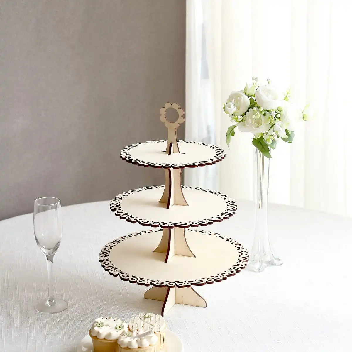 A three-level wooden cupcake or dessert stand with decorative laser-cut edges, displayed on a white table.