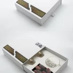 Sleek white drawer-style Ramadan box highlighted by golden Islamic motifs, opened to reveal elegant qahwa cups and fresh dates