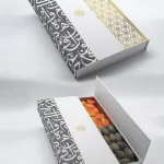 White cardboard gift box adorned with black Arabic calligraphy and a gold geometric accent strip, open to display dried fruits