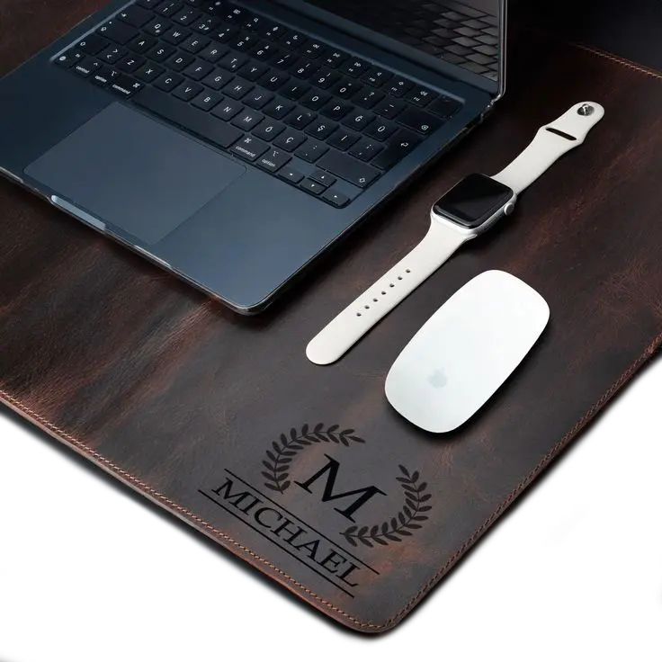 Brown leather desk mat engraved with a large ‘M’ and laurel wreath design, under a laptop and accessories 