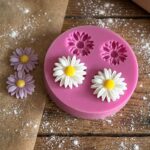 Pink food-grade silicone mold for making daisy-shaped chocolates and fondants, shown with finished white and purple candy flowers