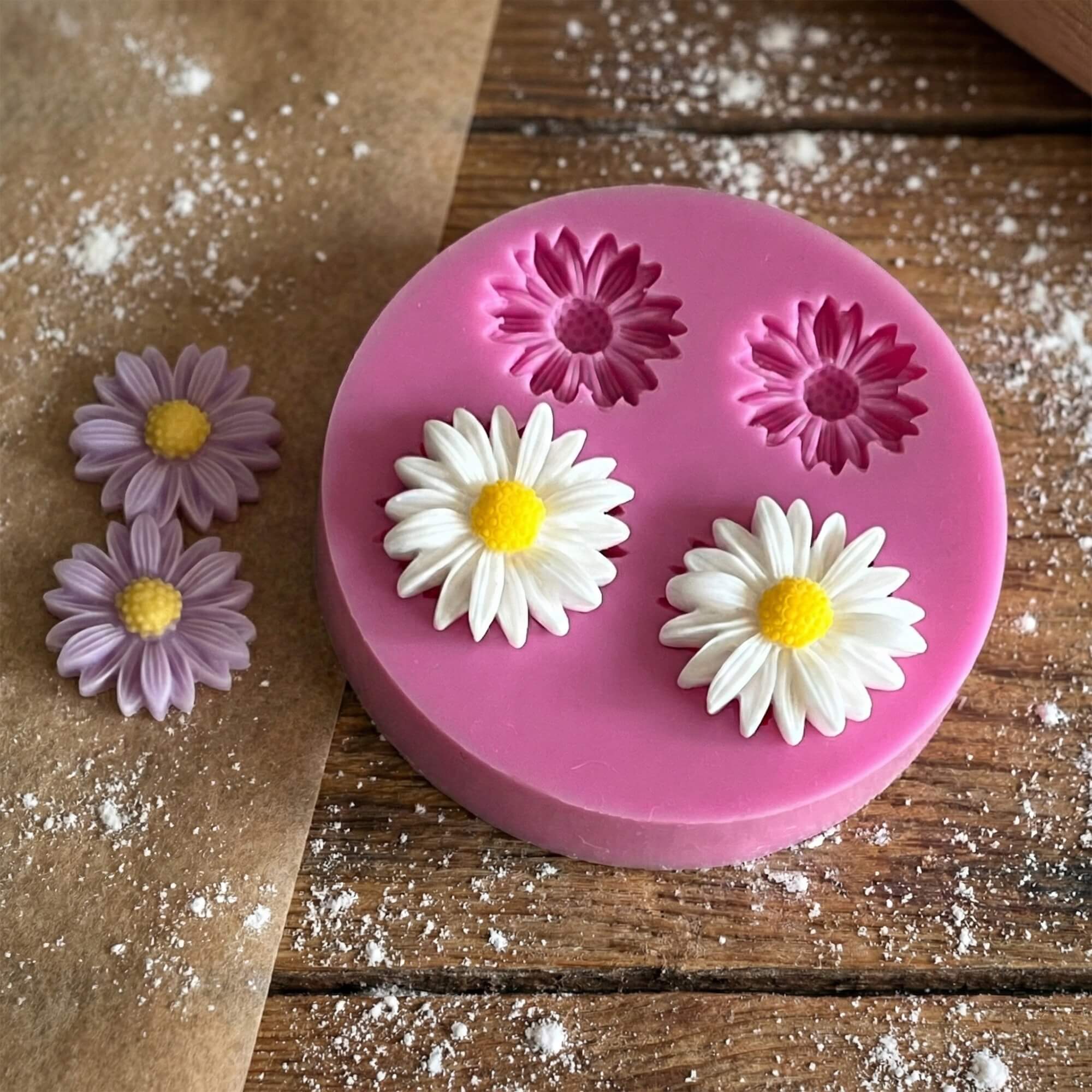 Pink food-grade silicone mold for making daisy-shaped chocolates and fondants, shown with finished white and purple candy flowers