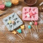 Grey and pink silicone lollipop molds with animal and egg designs, alongside finished colorful candies on sticks