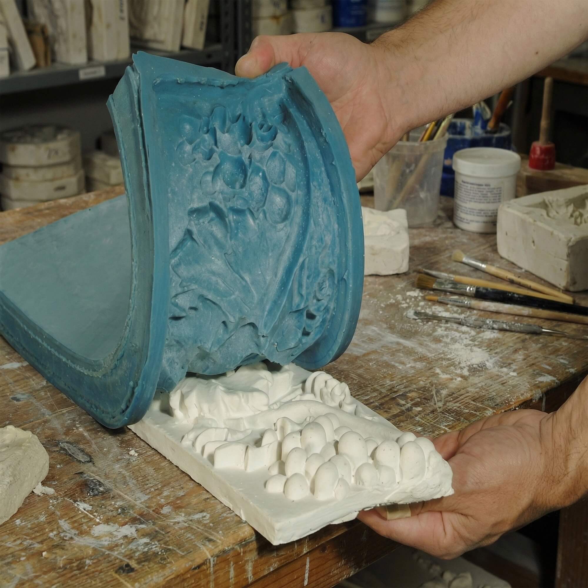 Hands peeling back a blue mold to reveal a detailed floral white gypsum relief panel.