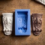 Blue silicone mold flanked by a white plaster cast and a bronze painted cast of an ornate architectural corbel.