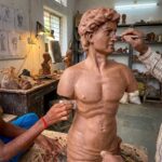 Two artisans using metal scraping tools to smooth and detail a large clay bust of a classical male figure in a production studio