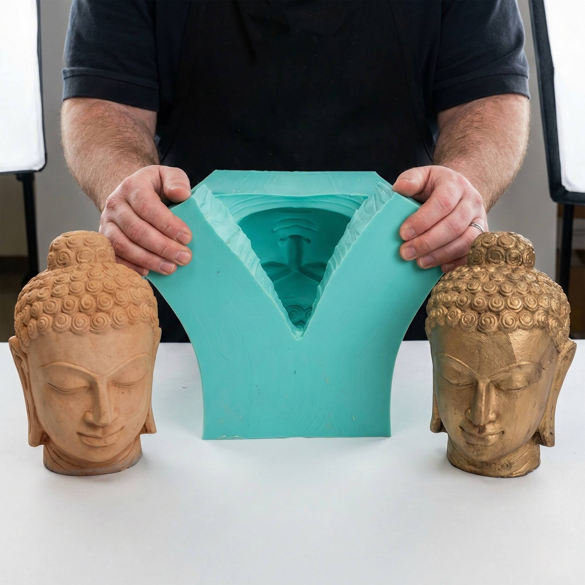 Artisan opening a large teal silicone mold to reveal a Buddha head cavity, with clay and gold cast models on the table.