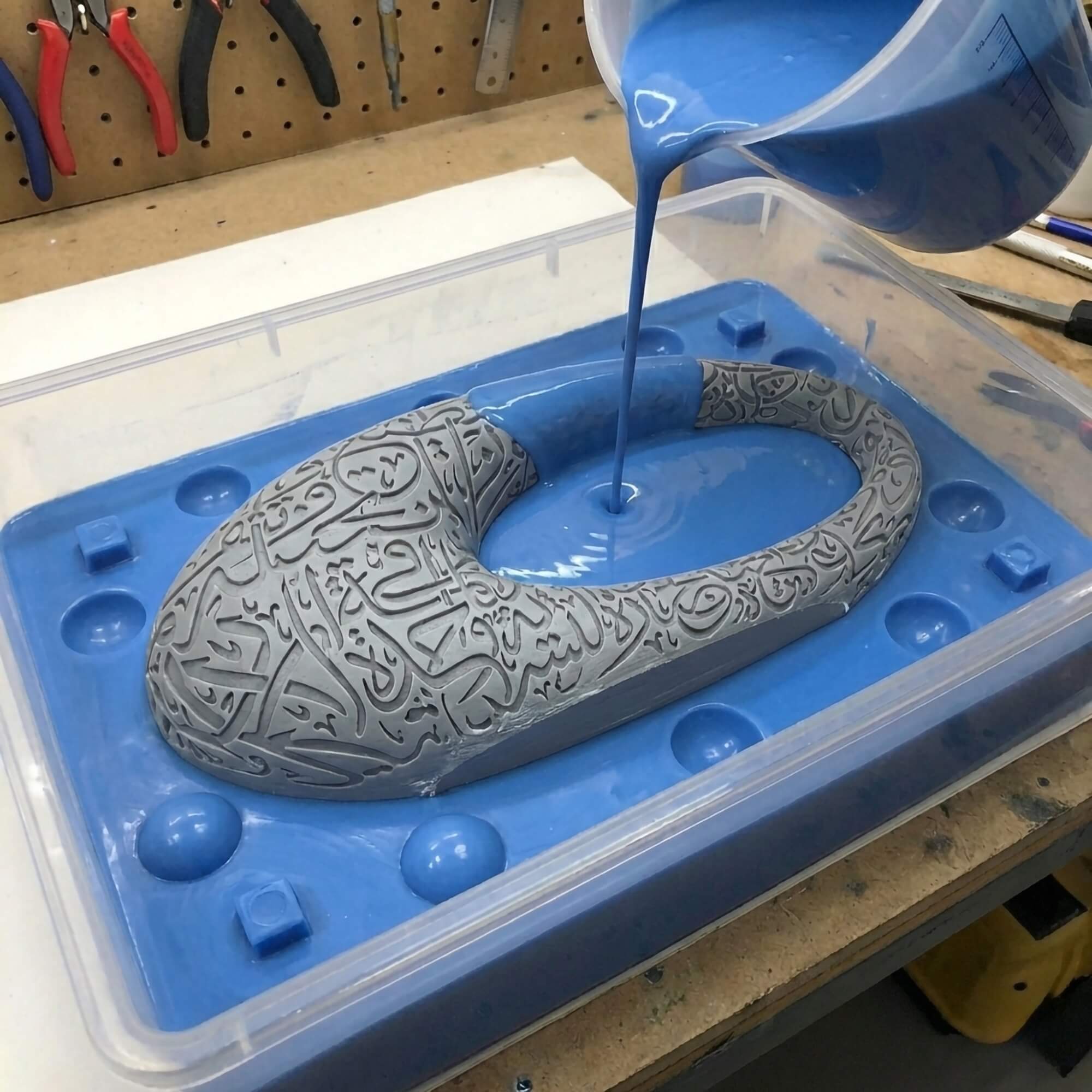 Pouring Liquid Silicone over Calligraphy Master Model Blue liquid silicone being poured over a master model featuring intricate Arabic calligraphy inside a plastic container. Custom mold making services
