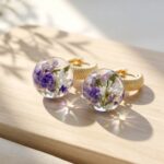 Pair of round clear casting resin earrings encapsulating purple and white dried flowers attached to textured gold hoops.