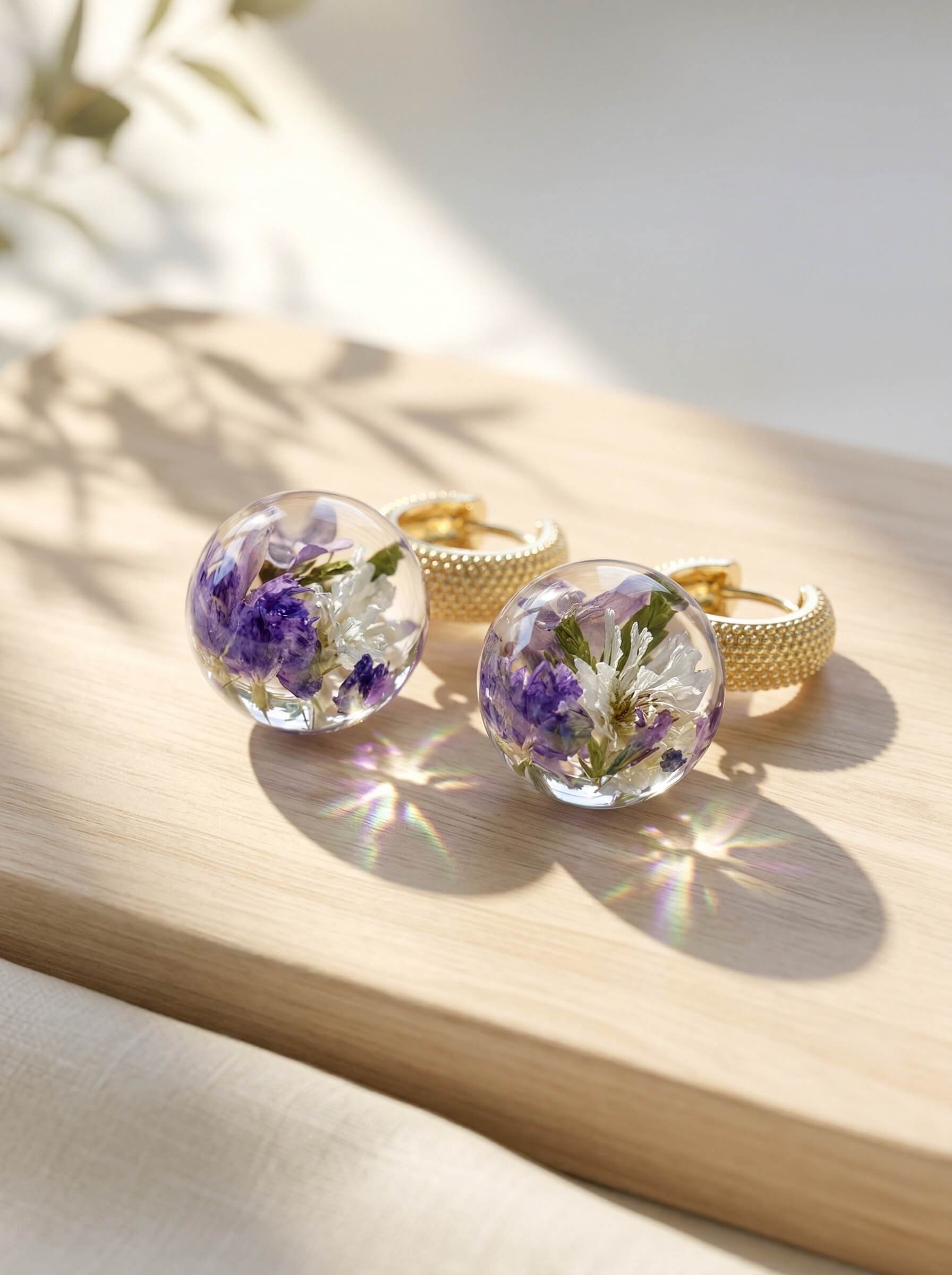 Pair of round clear casting resin earrings encapsulating purple and white dried flowers attached to textured gold hoops.