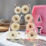 Clear casting resin letter A encapsulating whole fragile dandelions next to a pink silicone letter mold.