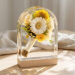 Arch-shaped clear epoxy resin block encapsulating a complete miniature wedding bouquet of yellow and white flowers tied with a white ribbon, resting on a light wood base.