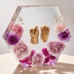 Clear hexagonal epoxy resin block encapsulating gold-painted baby foot and hand casts surrounded by pink and purple preserved roses