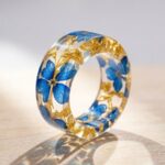 Solid clear epoxy resin ring encapsulating bright blue dried flowers and gold leaf casting colorful light refractions on a wooden surface.