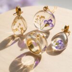 Clear epoxy resin botanical jewelry set including custom pendants and a gold-lined ring encapsulating purple and white dried flowers.