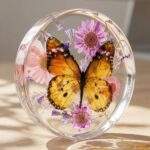 Round, polished clear epoxy resin paperweight encapsulating a large yellow butterfly surrounded by delicate pink and purple flowers.