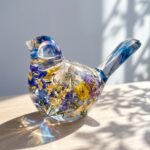 Clear epoxy resin bird figurine encapsulating dried blue and yellow flowers with gold flakes catching sunlight.
