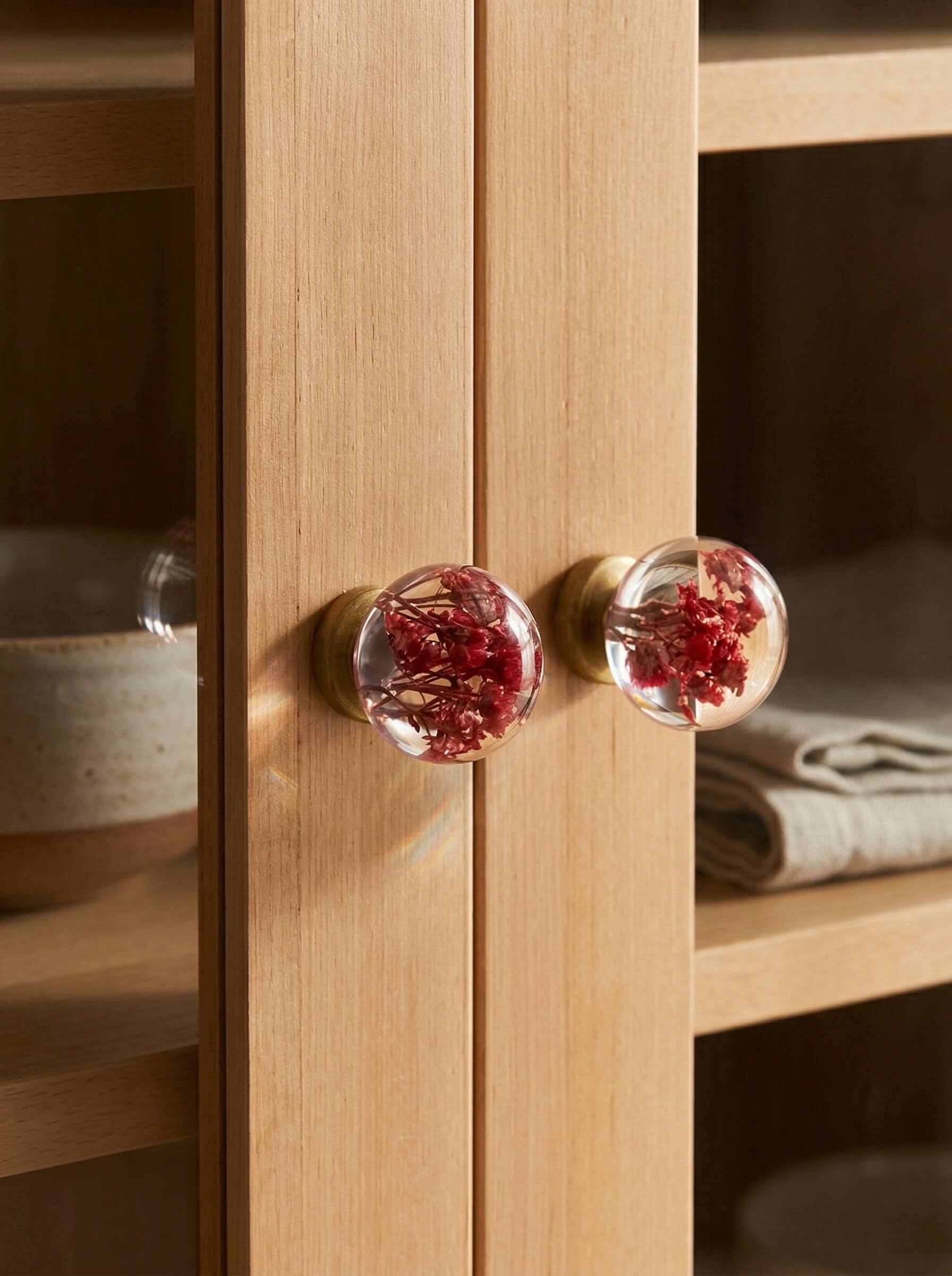 Round clear epoxy resin cabinet knobs encapsulating vibrant red dried flowers attached to a light wood cabinet door.