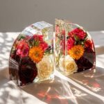 A pair of heavy, clear epoxy resin bookends encapsulating vibrant red, orange, and white roses sitting in bright sunlight.