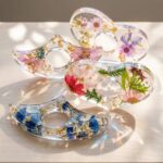 Clear epoxy resin ergonomic thumb page holders encapsulating tiny colorful flowers, leaves, and gold flakes on a wooden desk