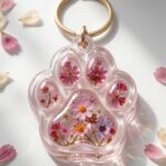Custom paw print shaped clear epoxy resin keychain encapsulating tiny pink and purple preserved flowers with gold hardware.