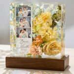 Clear epoxy resin block on a wooden base encapsulating a wedding photo strip, custom invitation text, and preserved yellow and blue flowers.