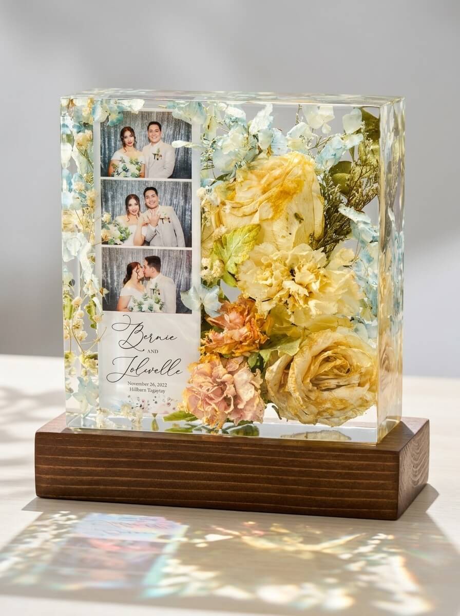 Clear epoxy resin block on a wooden base encapsulating a wedding photo strip, custom invitation text, and preserved yellow and blue flowers.