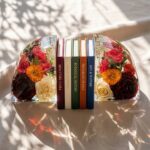 Clear epoxy resin floral bookends securely holding a row of colorful hardcover books on a sunlit table.
