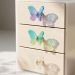 Three custom epoxy resin drawer knobs shaped like butterflies in translucent blue-purple, green-yellow, and clear finishes on a wooden dresser.