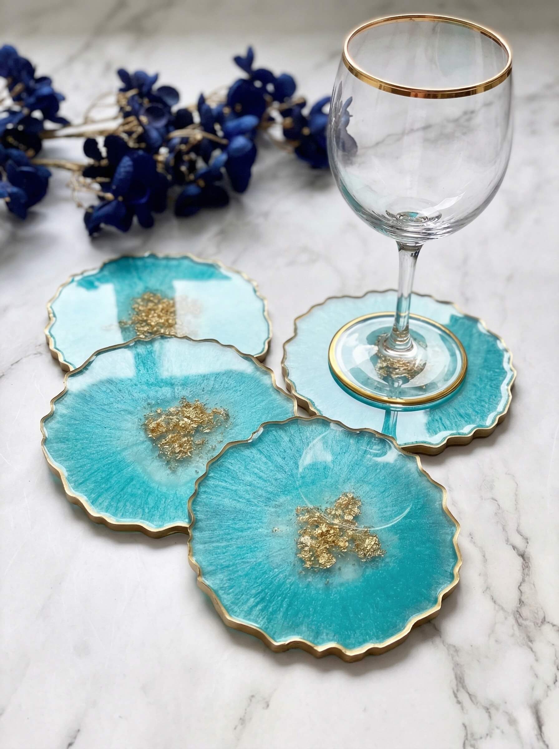 Set of three scalloped turquoise colored epoxy resin coasters featuring gold flake centers next to a wine glass.