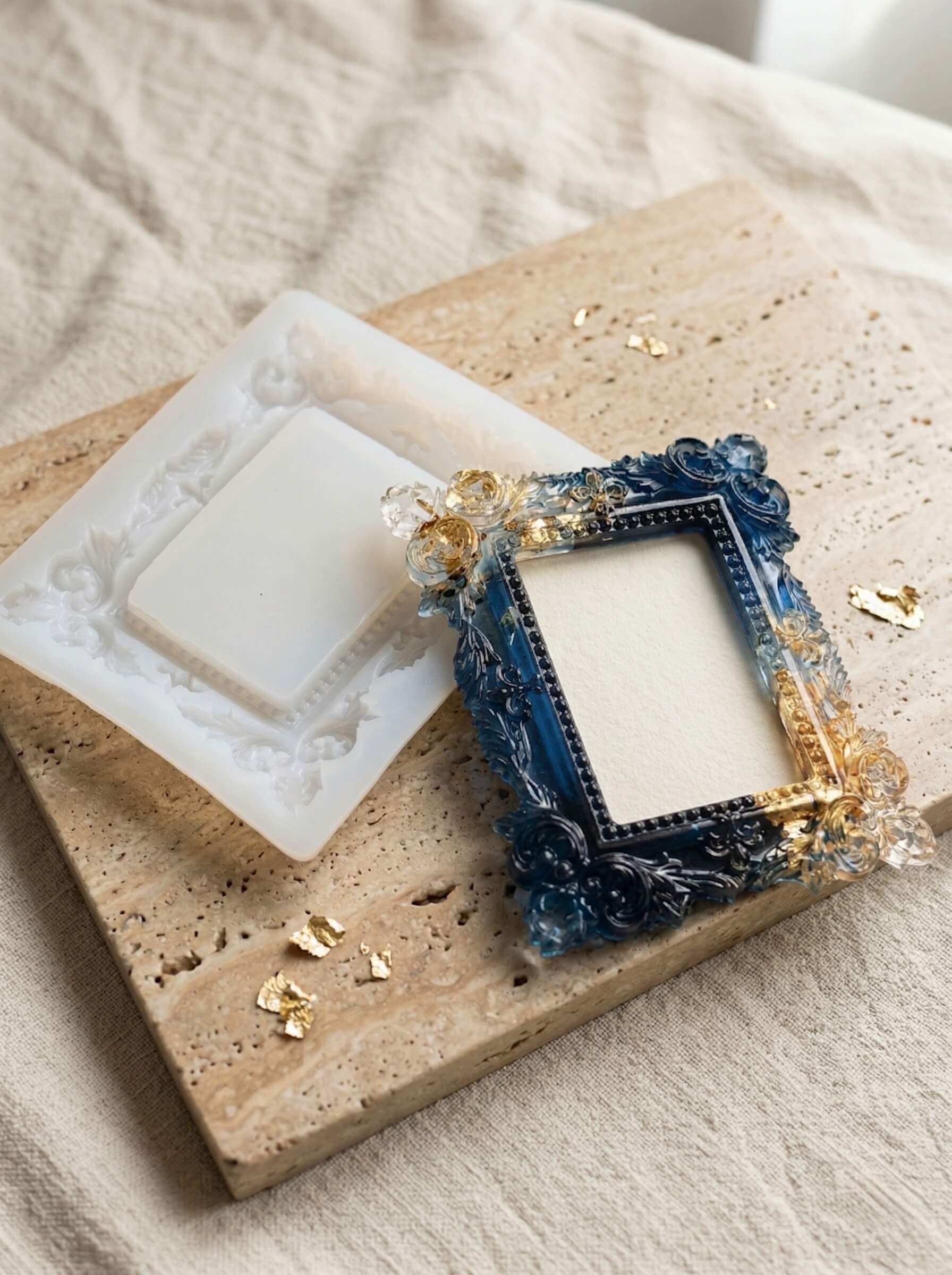 Ornate custom-cast colored epoxy resin photo frame in deep blue and gold resting next to its detailed silicone mold on a stone surface.