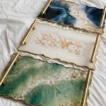 Three rectangular custom epoxy resin serving trays in dark blue, white, and emerald green with wavy gold edges and gold handles.