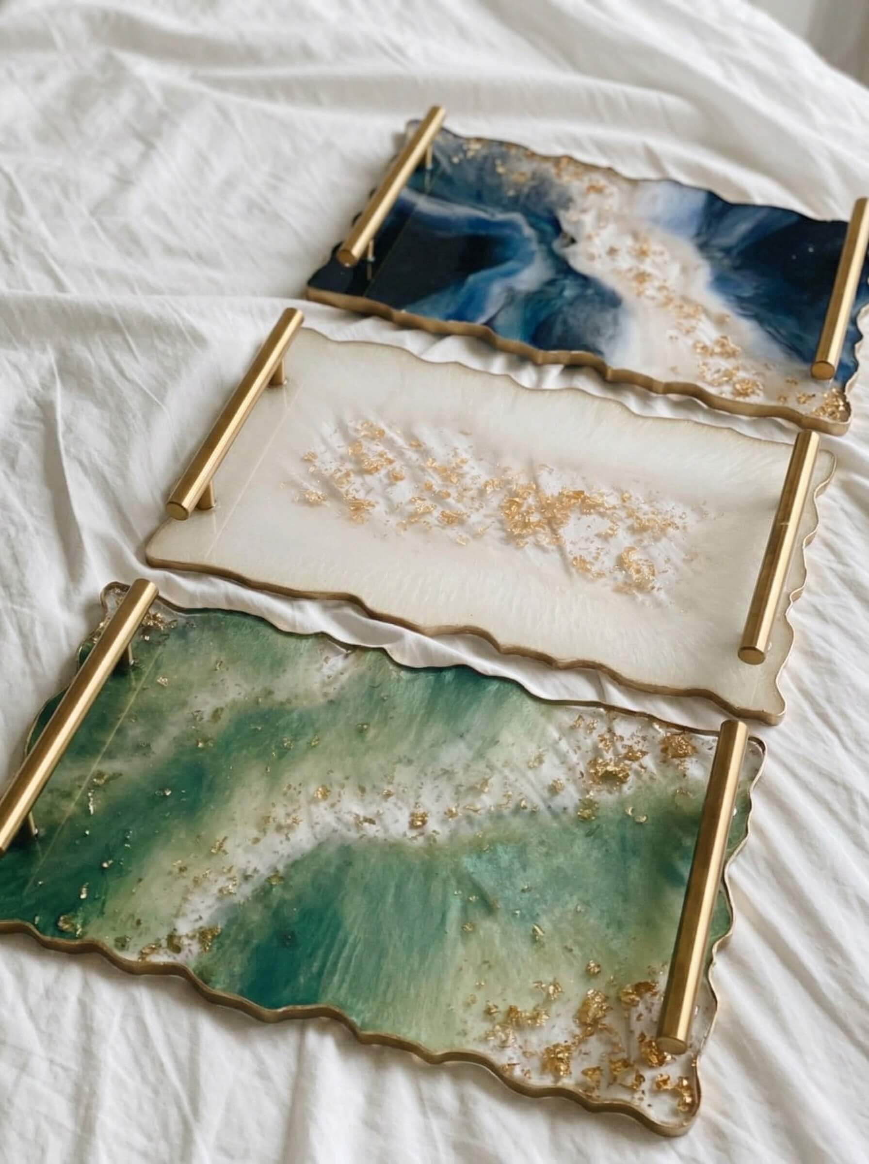 Three rectangular custom epoxy resin serving trays in dark blue, white, and emerald green with wavy gold edges and gold handles.
