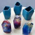 Three geometric colored epoxy resin crystals with gold flakes sitting next to custom blue silicone molds.