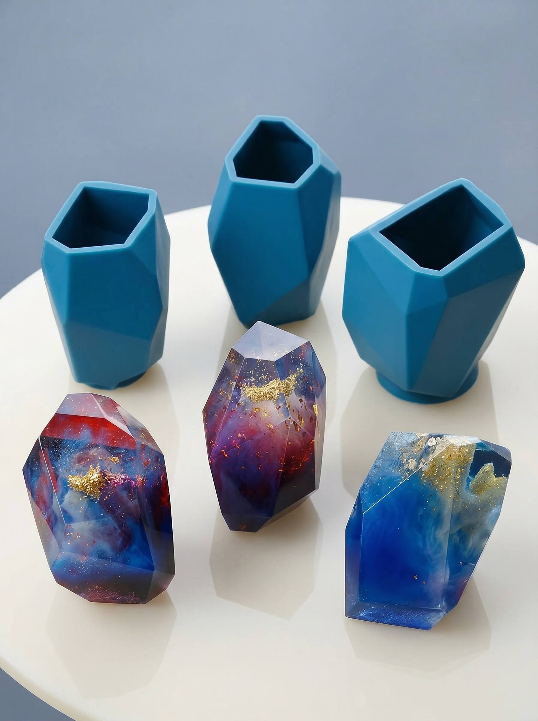 Three geometric colored epoxy resin crystals with gold flakes sitting next to custom blue silicone molds.