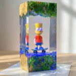 Tall clear epoxy resin block encapsulating a Bart Simpson action figure on a skateboard with textured terrain and gold leaf accents.