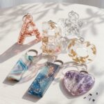 Assorted custom epoxy resin keychains in letter, heart, and star shapes encapsulating dried flowers, gold flakes, and swirling colors.