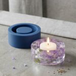 Clear epoxy resin candle holder with encapsulated purple flowers sitting next to its custom blue silicone mold.