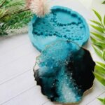 Textured geode style colored resin coaster in black, teal, and clear with gold edges next to a detailed silicone mold.