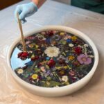A gloved hand using a wooden craft stick to carefully arrange a dense collection of dried flowers inside a large round silicone mold filled with liquid clear epoxy resin.