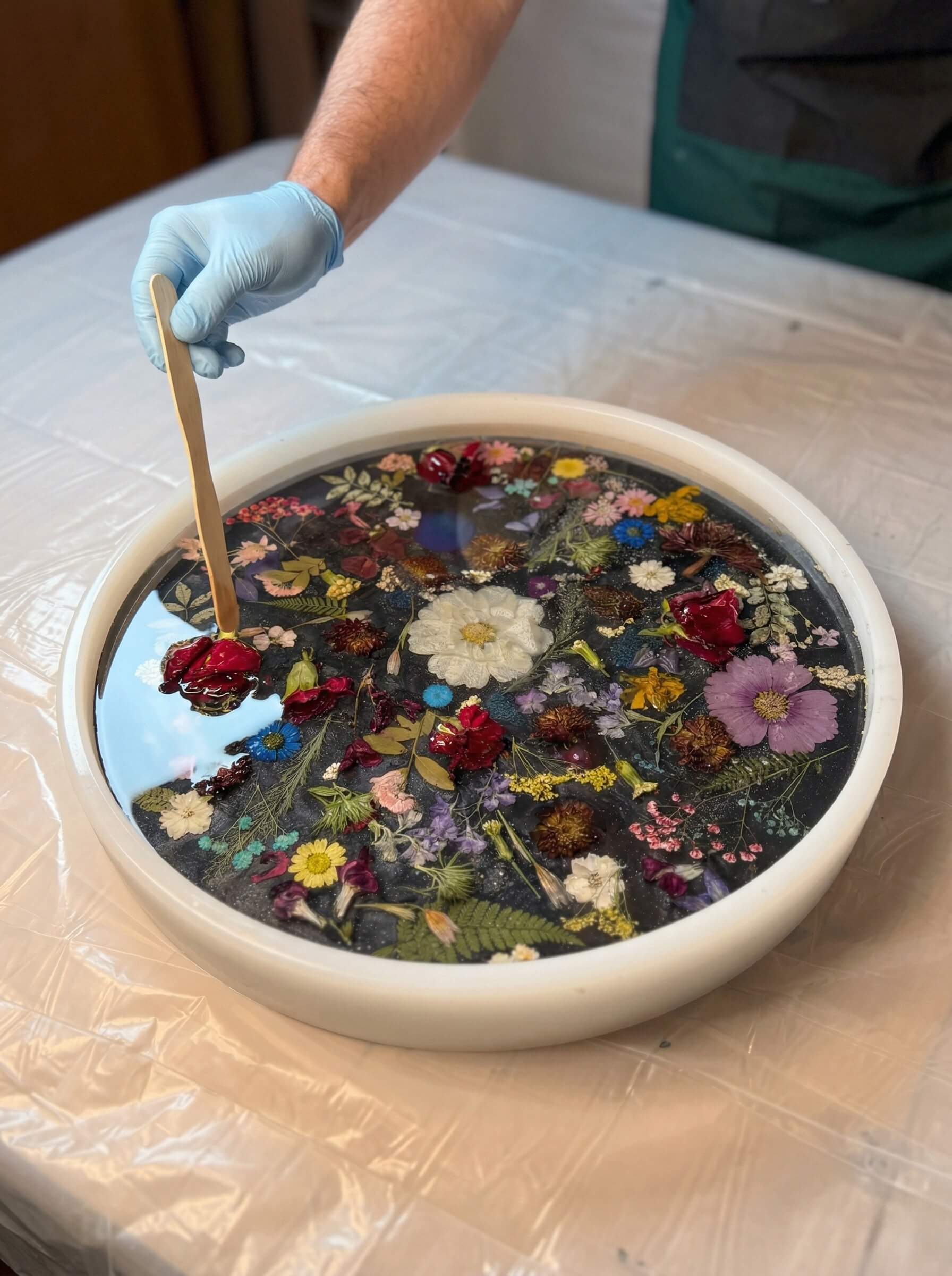A gloved hand using a wooden craft stick to carefully arrange a dense collection of dried flowers inside a large round silicone mold filled with liquid clear epoxy resin.